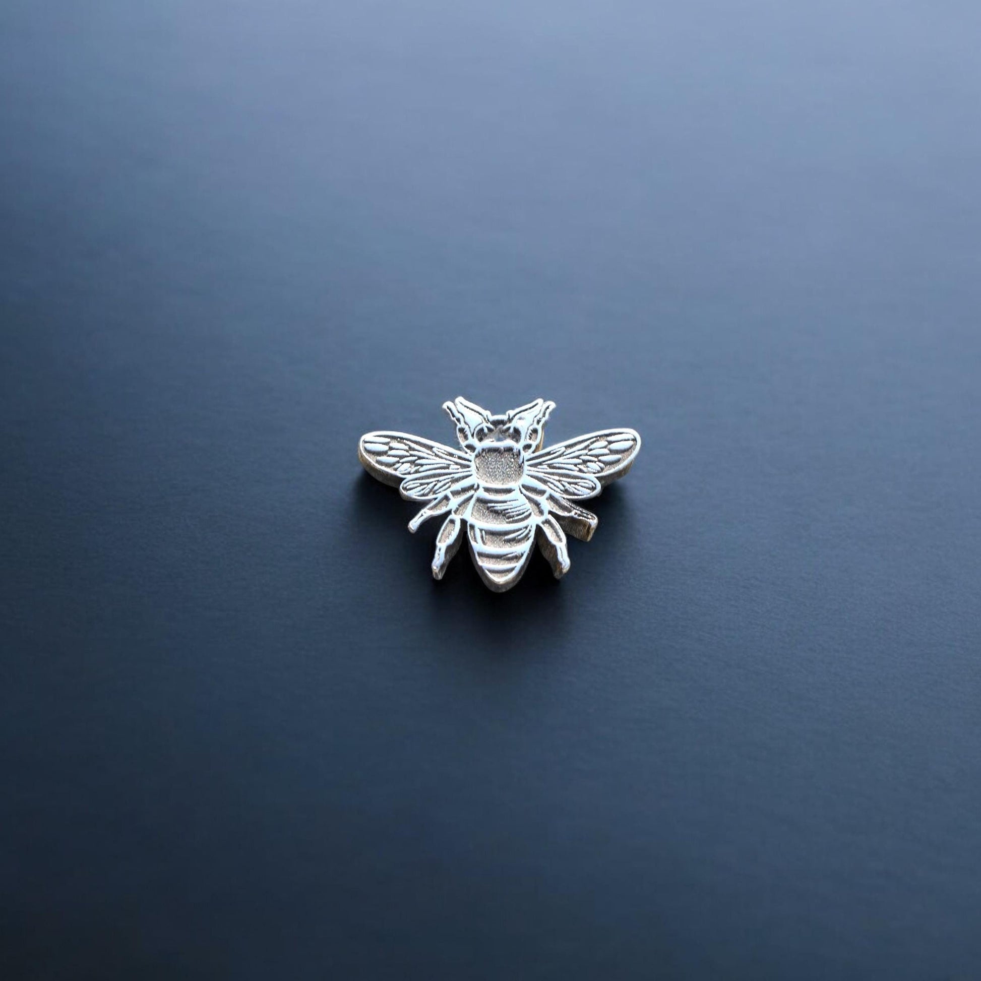 May include: A silver bee figurine with a white outline. The bee is facing forward with its wings spread out.