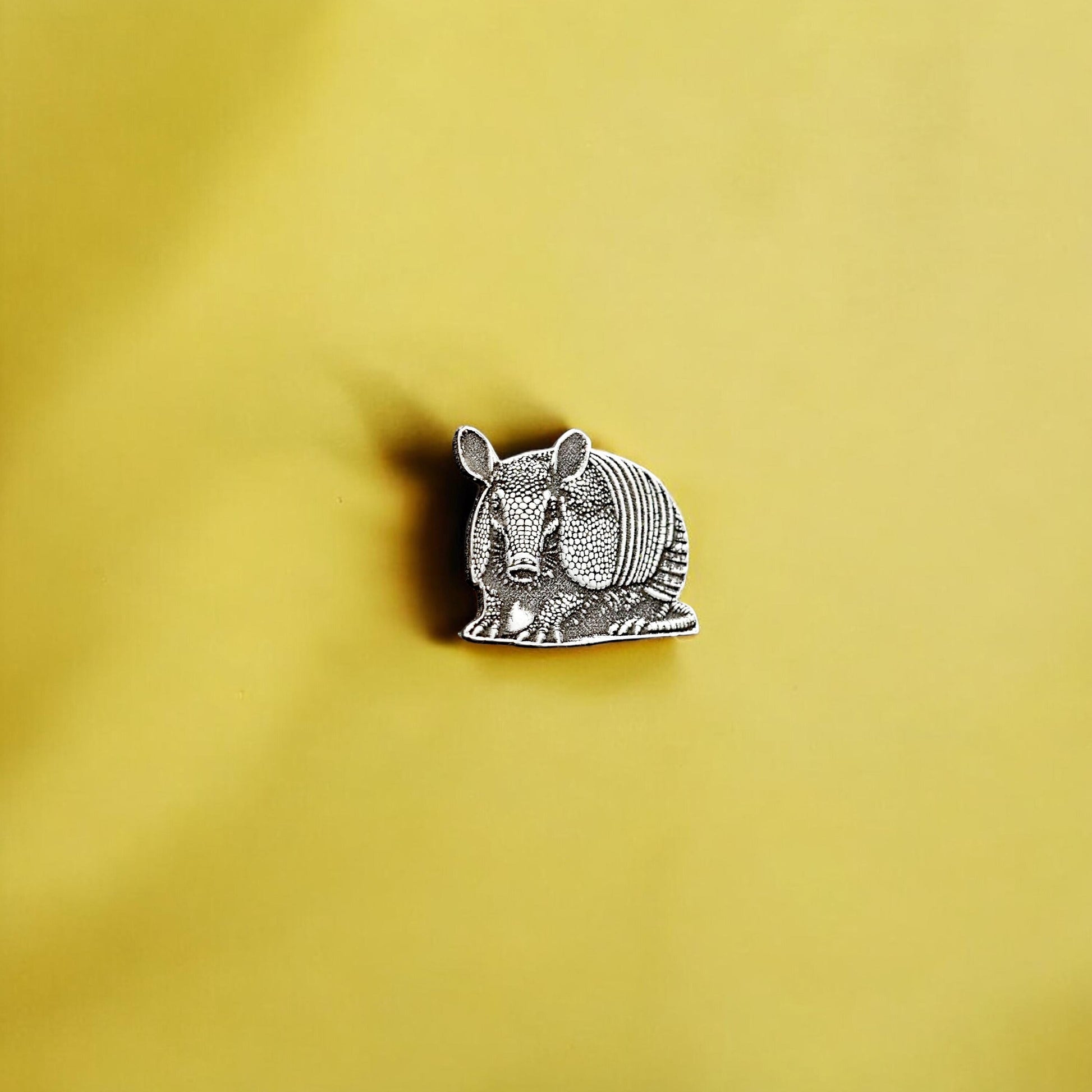 May include: A silver armadillo pin on a yellow background.