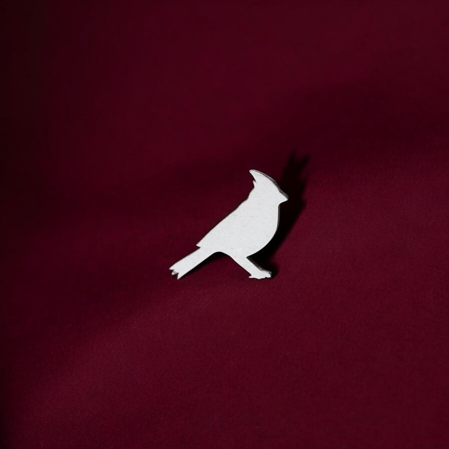 May include: White cardinal bird silhouette on a burgundy background. The bird is made of a solid white material and is positioned slightly off-center. The background is a deep burgundy color, providing a stark contrast to the white bird. The image is simple and elegant, suitable for various applications such as home decor, jewelry, or other craft projects. The bird's design is clean and modern, with smooth lines and a simple shape. The overall aesthetic is minimalist and sophisticated.