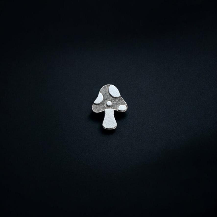 May include: Close-up view of a silver mushroom lapel pin. The mushroom cap is dark gray with small, lighter gray spots. The stem is a lighter shade of gray. The pin is displayed against a solid black background. The design is simple and minimalist, suitable for adding a touch of whimsy to clothing or bags.
